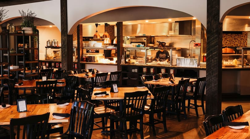 Rustic restaurant dining room with wooden tables, black chairs, and a visible open kitchen.