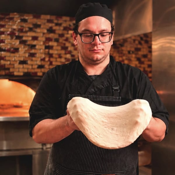 Chef in black uniform and glasses stretching pizza dough by a hot brick oven.