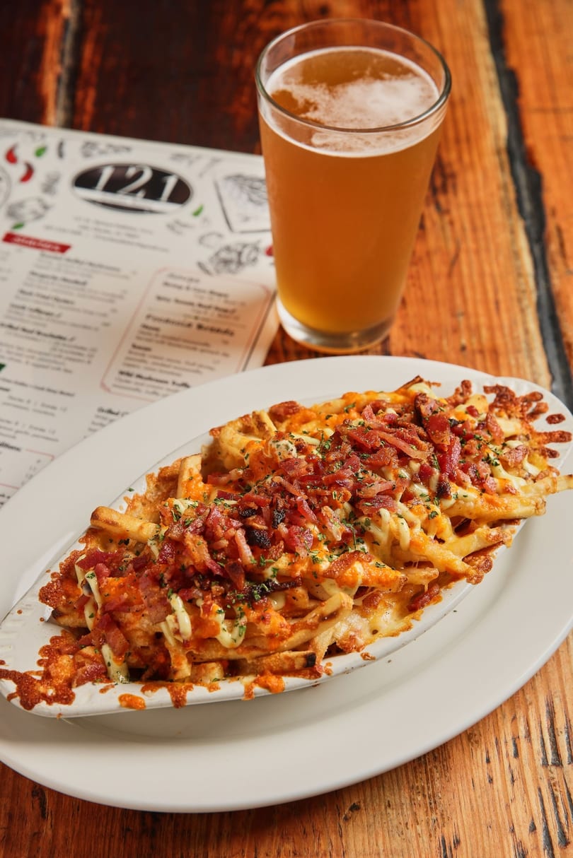A plate of cheesy bacon fries, a glass of beer, and a menu on a wooden table.