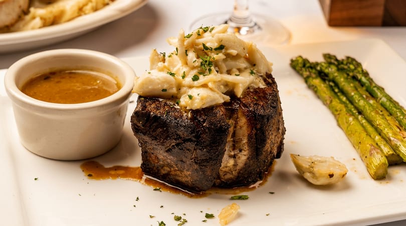 Steak topped with crab meat, served with asparagus, a side of sauce, and roasted garlic.