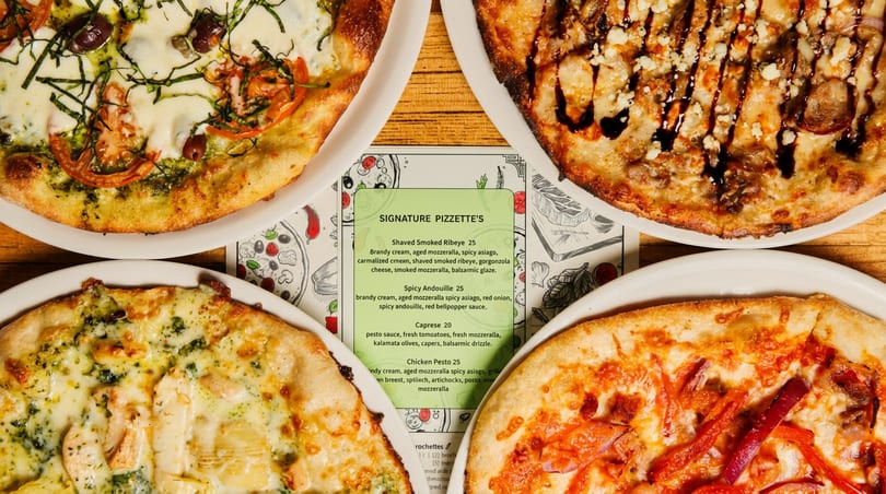Four diverse pizzas surround a "Signature Pizzette's" menu card on a wooden table.