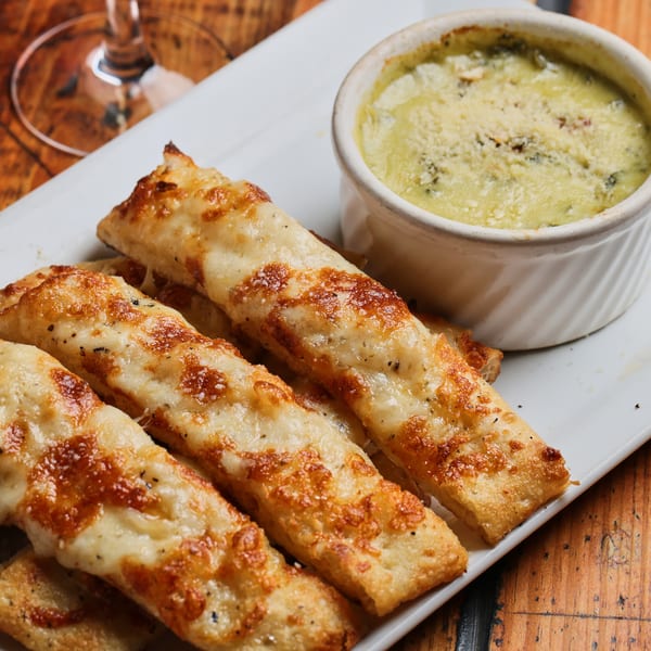 Golden cheesy breadsticks served on a white platter with a ramekin of creamy herb dip on a wooden table.