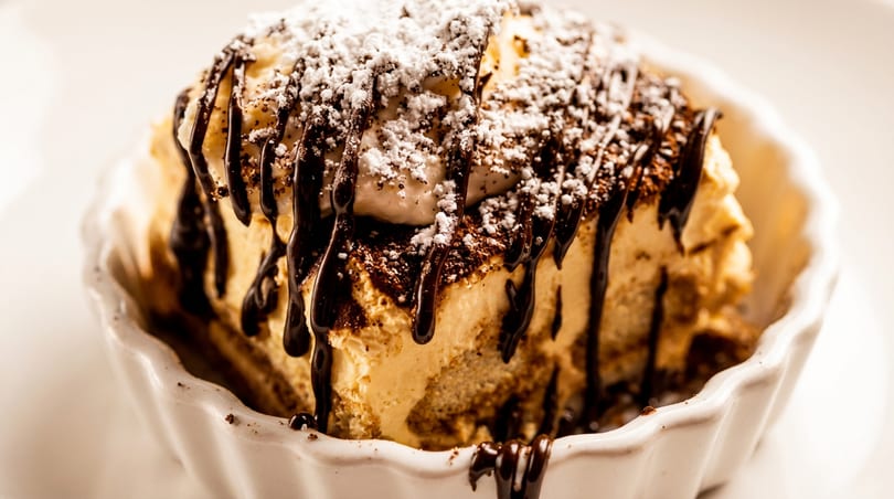 Tiramisu dessert with chocolate drizzle, powdered sugar, and cocoa in a white fluted dish.