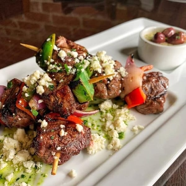 Two grilled meat and vegetable skewers with feta cheese, green sauce, and a side of olives.