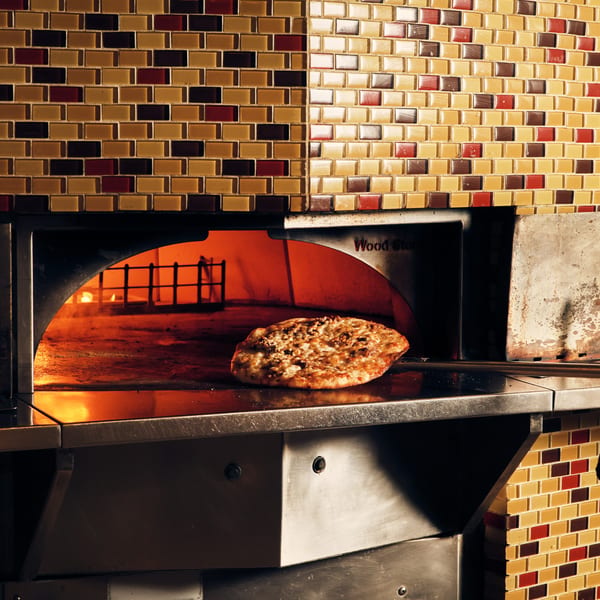 A pizza with melted cheese and toppings baking inside a wood-fired oven with a tiled exterior.