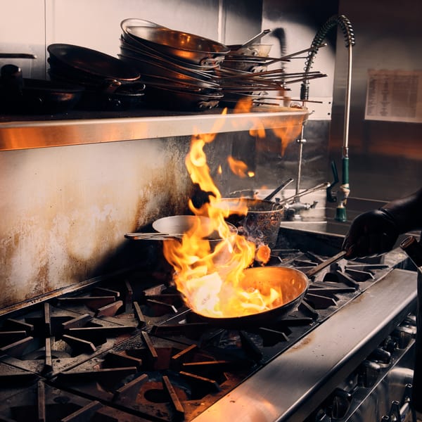 Chef flambéing food in a pan on a commercial kitchen stove, with stacked pans nearby.