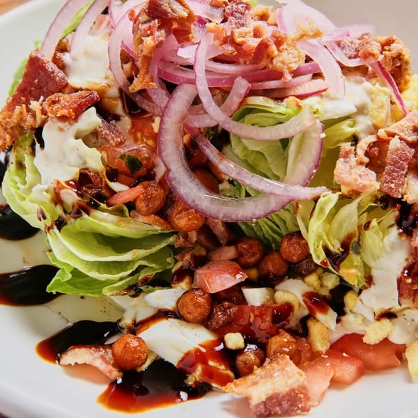 Wedge salad with bacon, red onion, tomatoes, and dressing on a white plate.