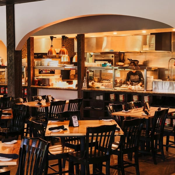 Inviting restaurant dining area with wooden tables, black chairs, and an open kitchen visible through arches.