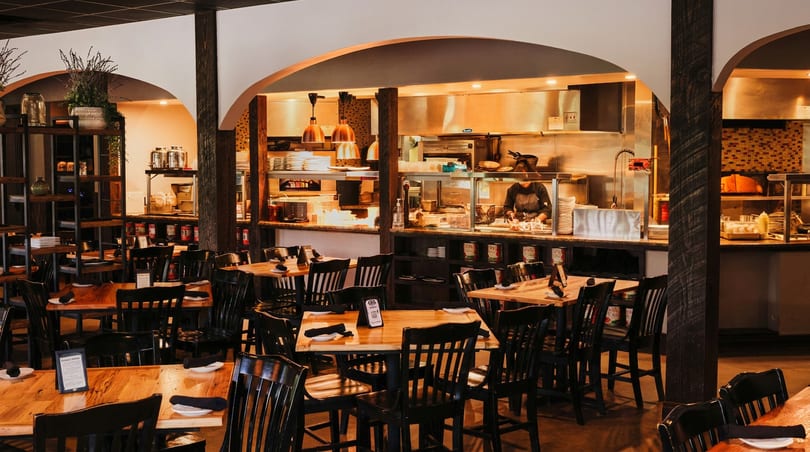 Warm restaurant interior with dining tables and an open kitchen where a chef prepares food.