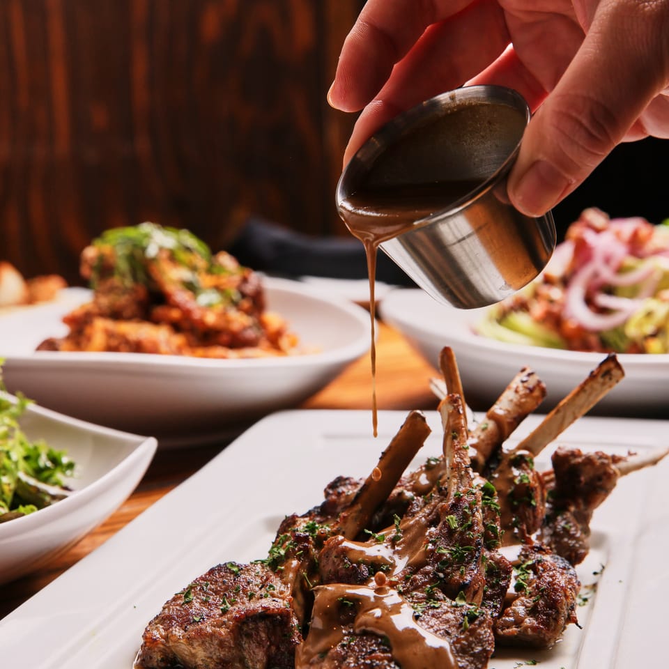Hand pours brown sauce from a metal pitcher over grilled steak medallions garnished with herbs on a white platter.