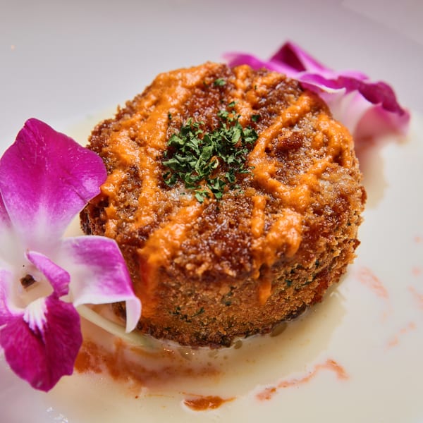 Crispy crab cake with orange sauce, parsley, and two purple orchids on a white plate.