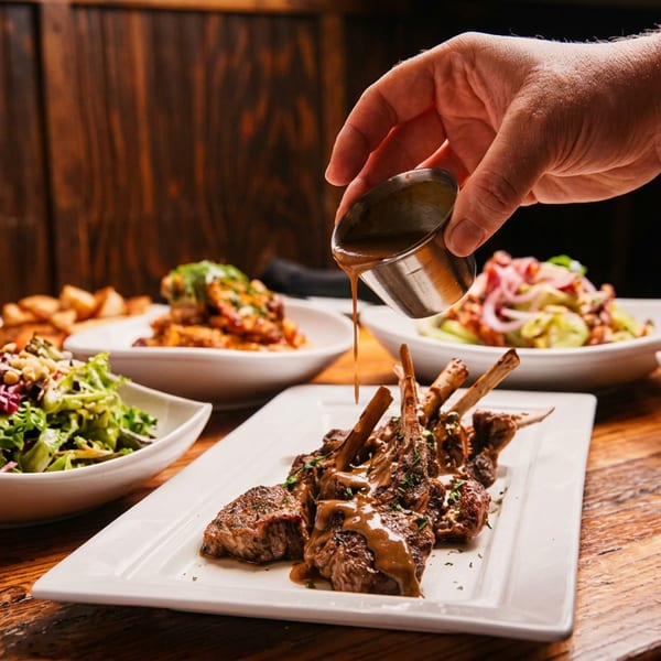A hand pours sauce over a plate of lamb chops, surrounded by salads and potatoes on a wooden table.
