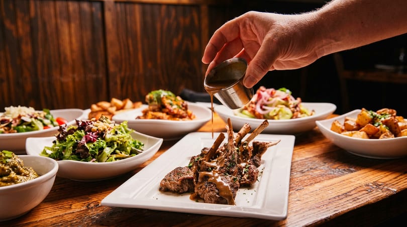 Hand drizzles sauce on lamb chops, surrounded by salads and side dishes on a wooden table.