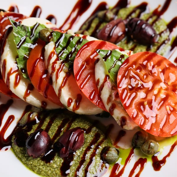 Caprese salad with sliced tomatoes, mozzarella, basil, balsamic glaze, pesto, olives, and capers.