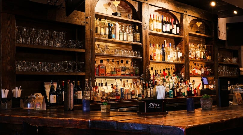 Rustic wooden bar counter with numerous liquor bottles and glassware on illuminated shelves.