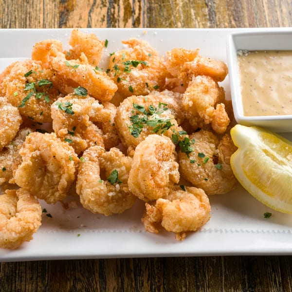 Crispy fried shrimp with parmesan, parsley, a lemon wedge, and dipping sauce on a white plate.
