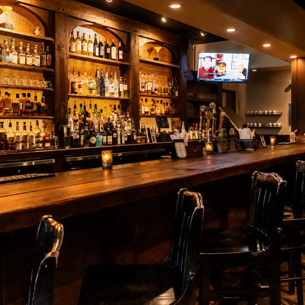 Warmly lit restaurant bar with a long wooden counter, black chairs, glowing candles, and shelves stocked with liquor bottles.