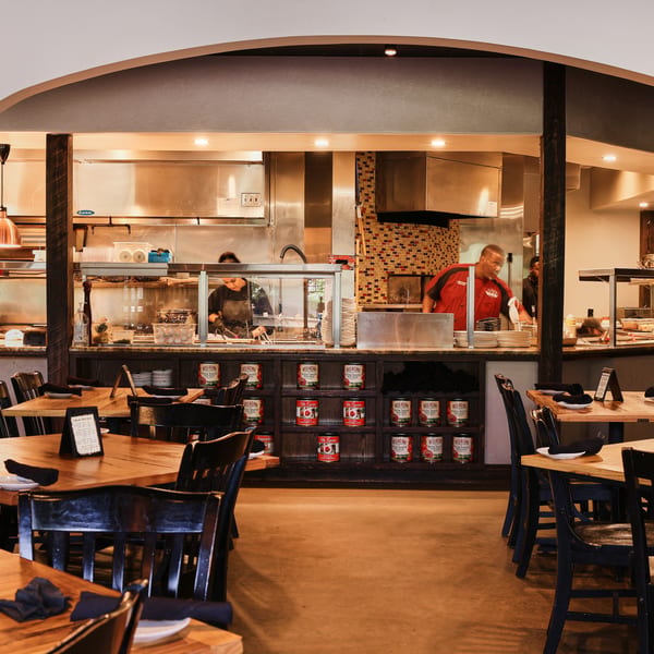 Restaurant dining room with wooden tables and chairs, and an open kitchen with chefs working.