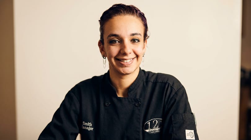 Smiling woman in a black chef's jacket with "Smith Manager" and "121 Artisan Bistro" logos.