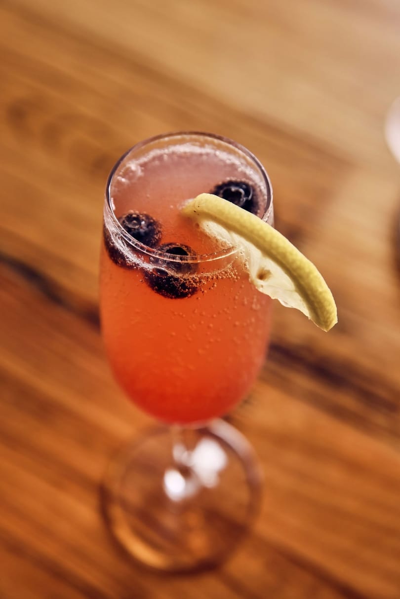 Fizzy reddish-orange cocktail with blueberries and lemon slice in a stemmed glass on wood.