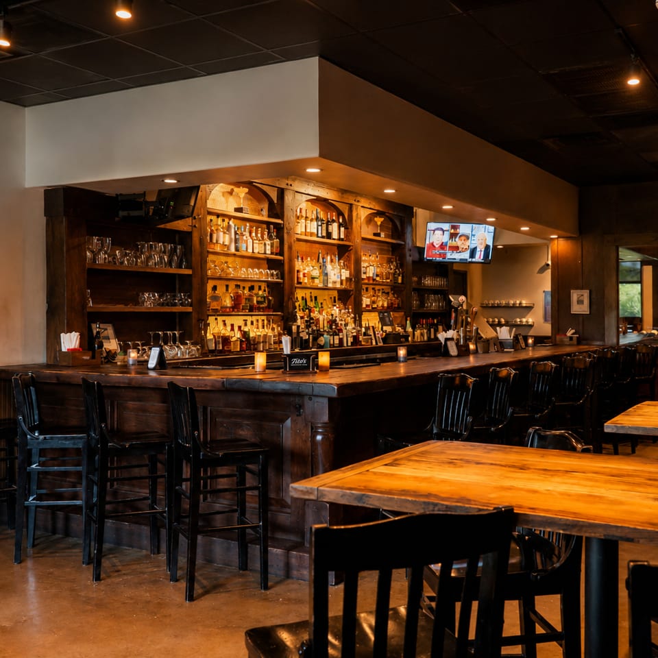 Warmly lit restaurant interior featuring a fully stocked wooden bar, black chairs, wooden tables, candles, and a wall-mounted television.
