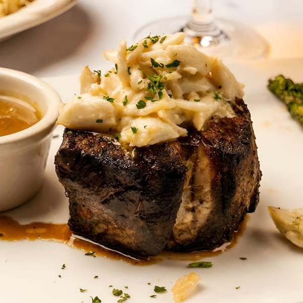 Seared steak topped with crab meat, served with a side of sauce and grilled asparagus.