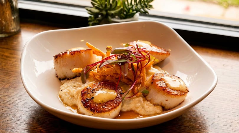 Plate of seared scallops on creamy grits, topped with crispy onions and red microgreens.