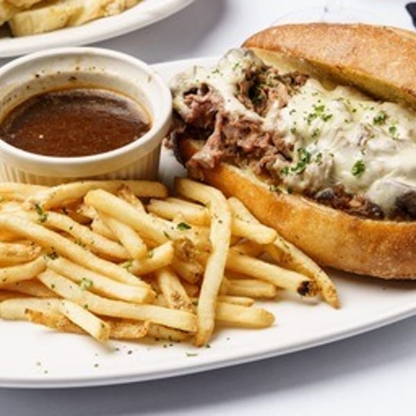 A French dip sandwich with melted cheese, a side of fries, and au jus on a white plate.