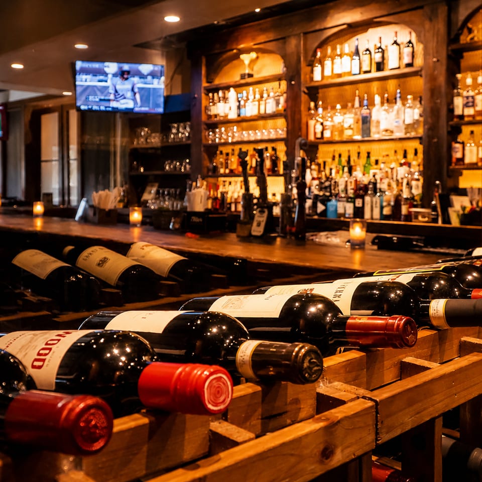 Rows of wine bottles in wooden racks overlook a warmly lit restaurant bar stocked with liquor and glasses.
