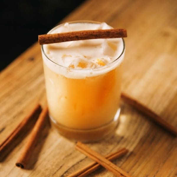 Iced cocktail with foam and a cinnamon stick on a wooden table with scattered cinnamon.