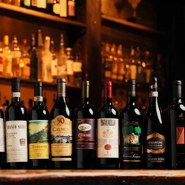A row of various red wine bottles on a wooden bar with blurred bottles in the background.