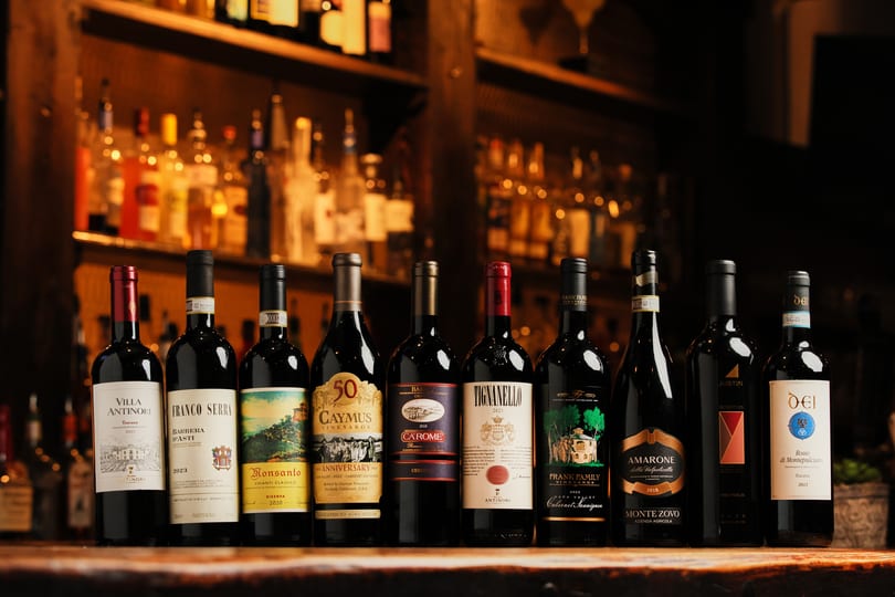 A row of various red wine bottles on a wooden bar with blurred bottles in the background.