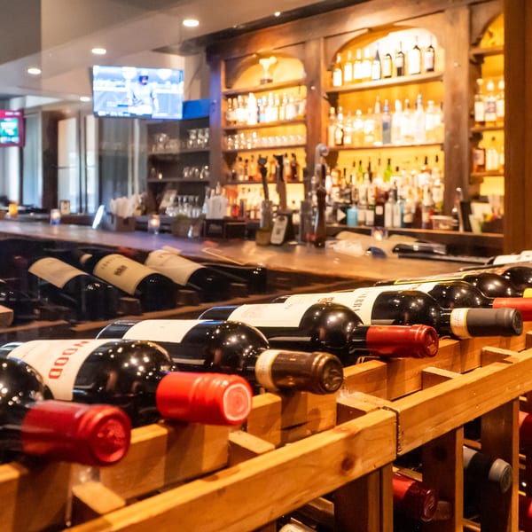 Wine bottles in wooden racks in the foreground, with a dimly lit bar and liquor shelves behind.