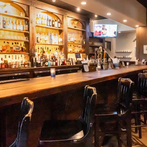 A dimly lit bar with a long wooden counter, bar stools, and shelves filled with liquor bottles.