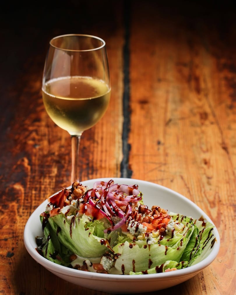 Wedge salad with blue cheese, tomatoes, red onion, and balsamic glaze, beside a glass of white wine.