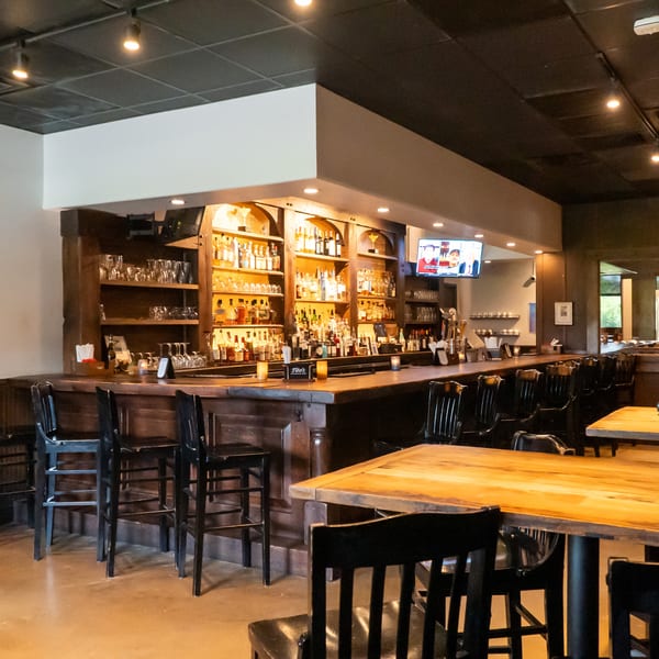 A cozy bar area with a fully stocked wooden bar, black bar stools, and dining tables.