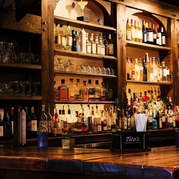 Warmly lit wooden bar counter with shelves full of liquor bottles and glassware.