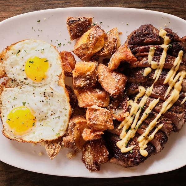 Sliced steak with creamy sauce, sunny-side up eggs, and parmesan roasted potatoes on a white plate.