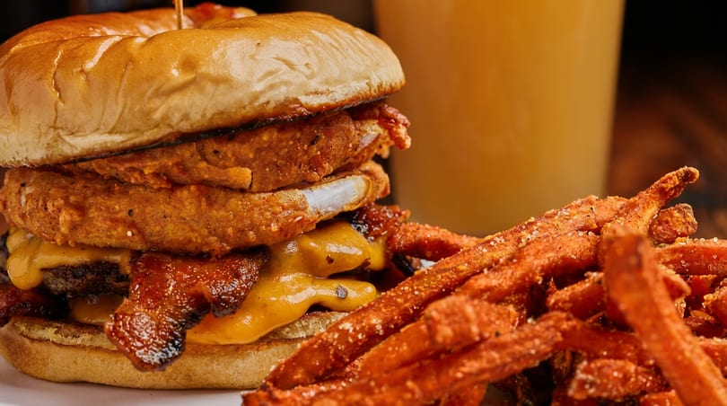 Loaded burger with bacon, melted cheese, and onion rings, served with sweet potato fries and a drink.
