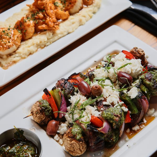 Two plates of food, one with scallops and shrimp over mashed potatoes, the other with grilled vegetable skewers.