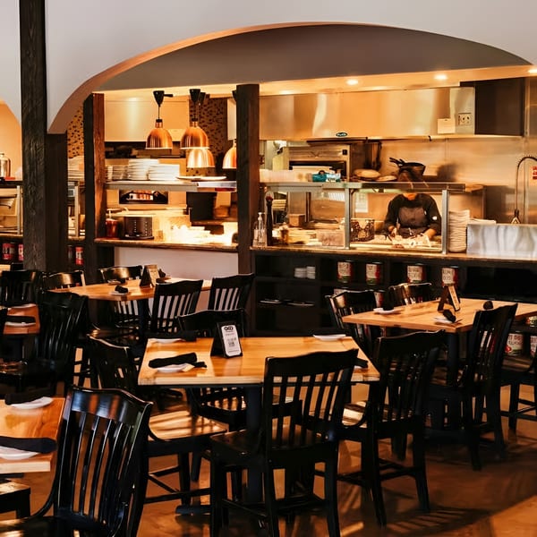 Cozy restaurant interior with wooden tables, black chairs, and an open kitchen where a chef works.