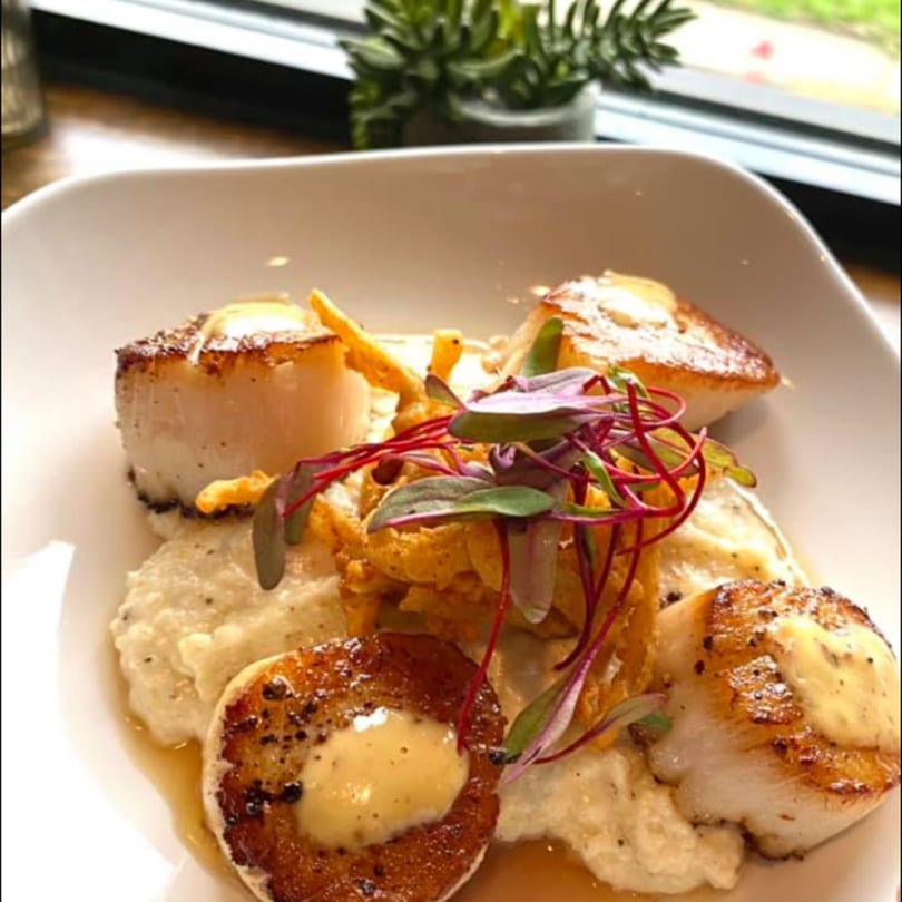 Seared scallops with creamy grits, crispy fried onions, and microgreens on a white plate.