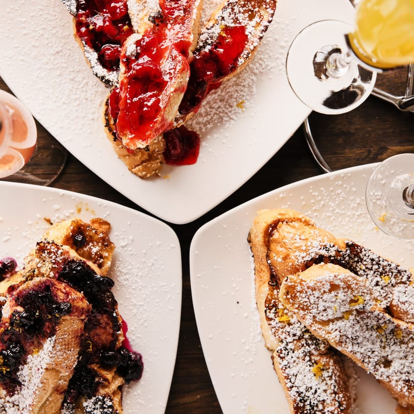 Three plates of French toast with powdered sugar and fruit toppings, with drinks on a dark table.