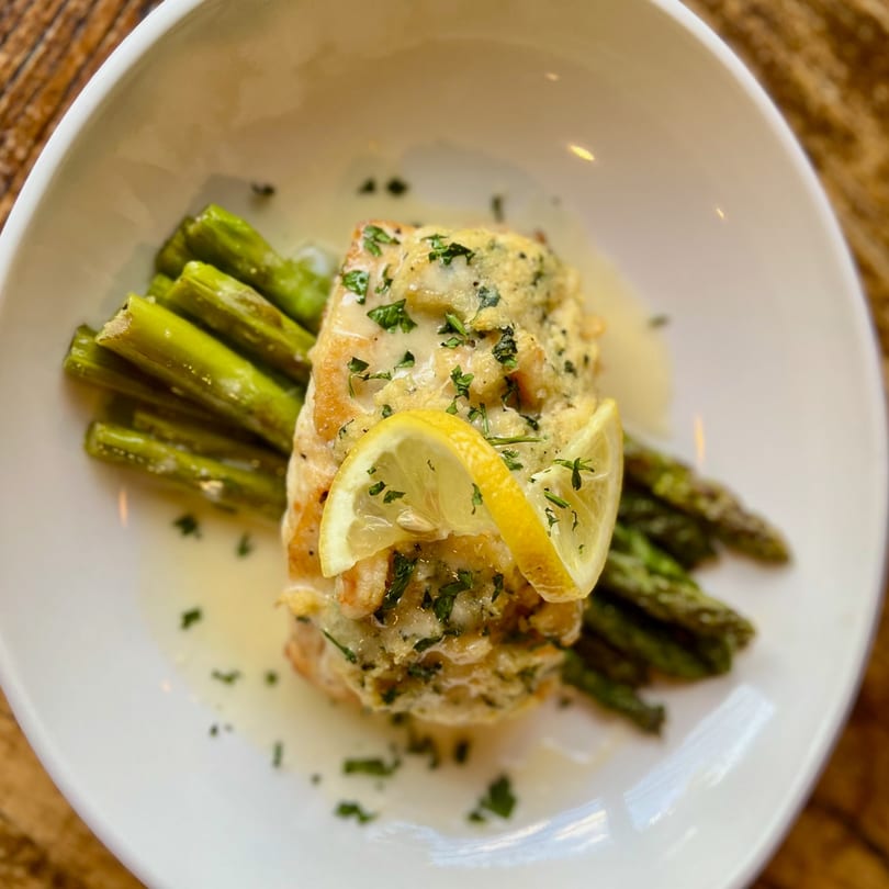 Baked fish with lemon slices and parsley, served with asparagus in a white bowl.