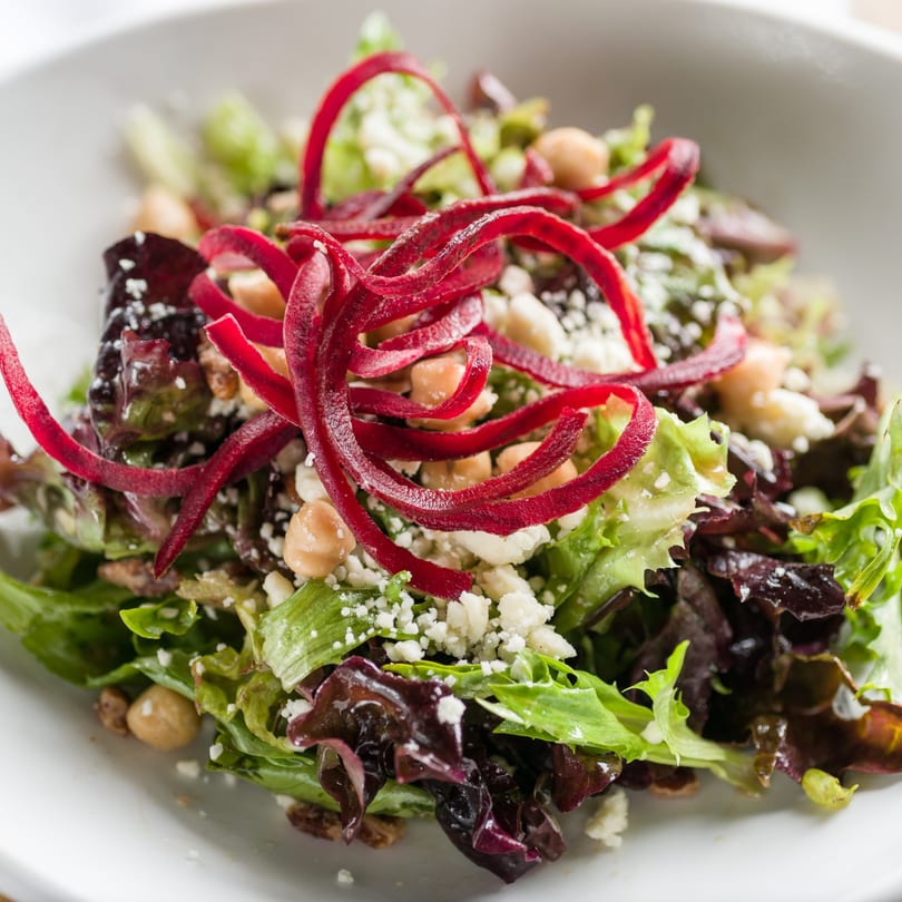 Close-up of a fresh salad with mixed greens, chickpeas, crumbled cheese, and red beet ribbons.