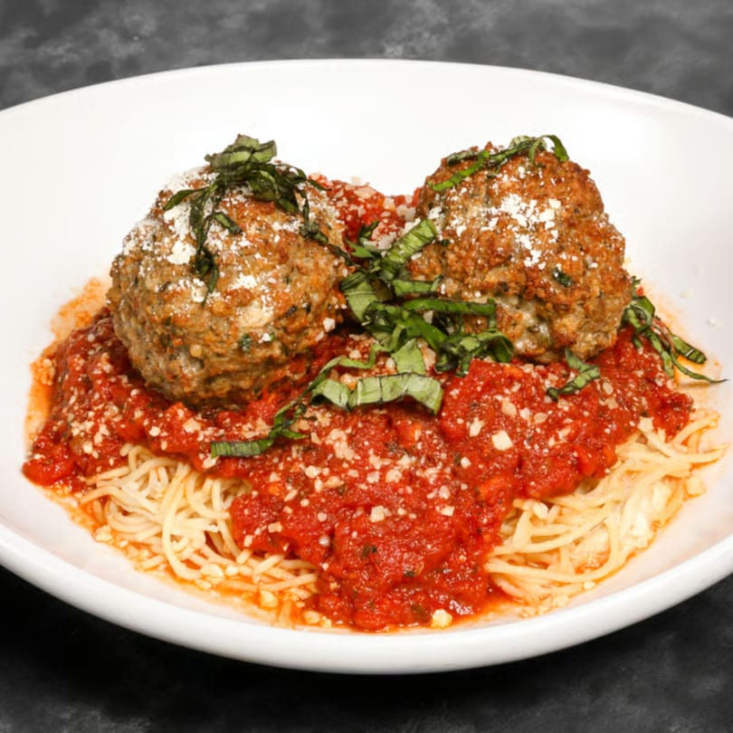 Spaghetti with two large meatballs, marinara sauce, grated cheese, and fresh basil in a white bowl.