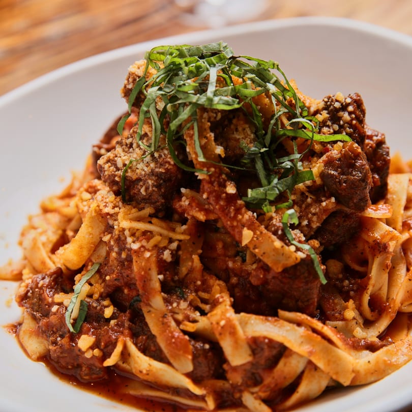Close-up of tagliatelle pasta with rich red sauce, meatballs, grated cheese, and fresh basil.