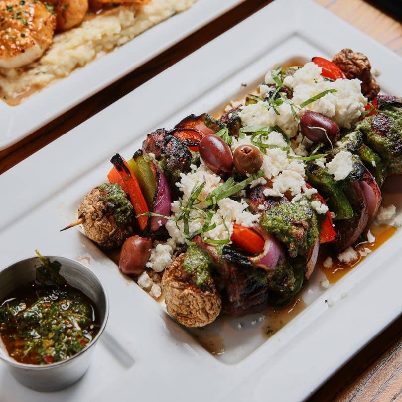 Grilled vegetable and meat skewers topped with feta, olives, and green sauce, with a side of sauce.