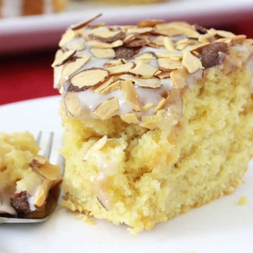 A slice of almond cake with white frosting and toasted almond slivers on a white plate.